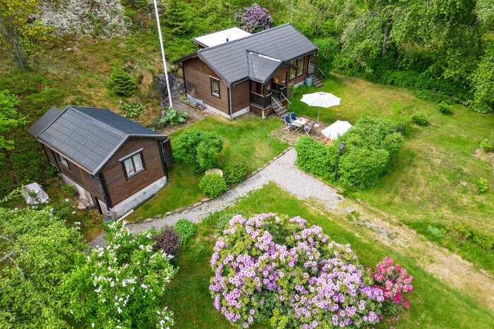 Luftfoto av ett 1-plans trähus med svart tak, beläget i en lummig trädgård med grusgång och färgglada buskar.