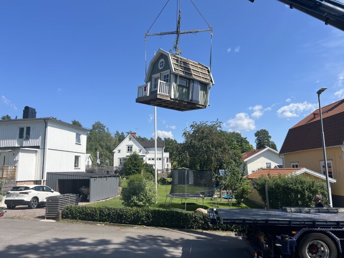 Lekstuga hänger i luften lyft av en kran, omgiven av hus och grönska, under en blå himmel.