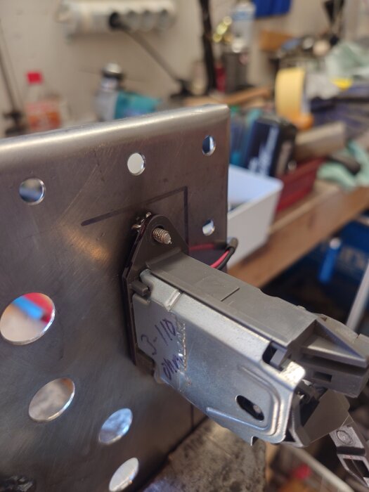 Detaljbild av ett metallfäste med rostfri skruv och elektrisk komponent i en verkstadsmiljö.