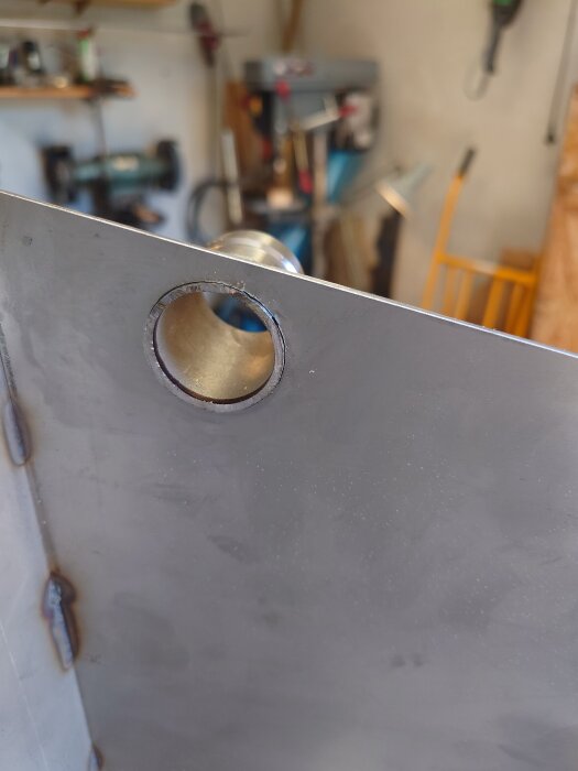 Metallplåt med rund öppning och svetsade fogar syns i en verkstadsmiljö med diffust bakgrund.
