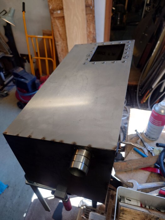 Rektangulär metalltank under konstruktion med en svetsad kant och en rördel, omgiven av verktyg i en verkstadsmiljö.