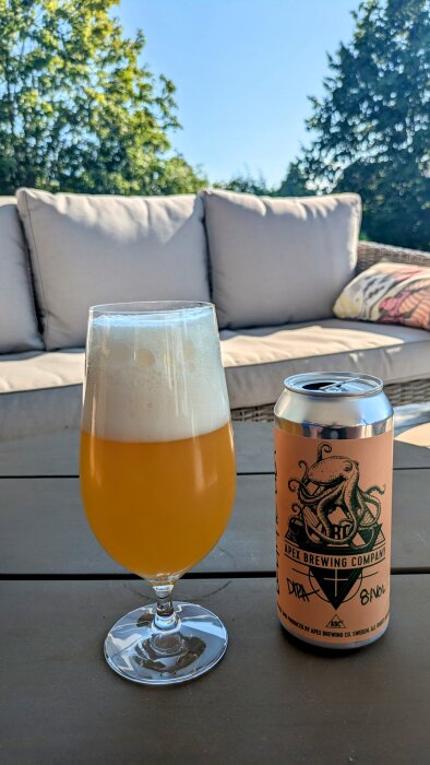 Ett glas orange öl med skum bredvid en ölburk från Apex Brewing Company på ett bord framför en grå soffa utomhus.