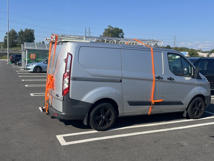 Grå skåpbil med stege fäst på taket med orange spännband, parkerad på en asfaltsparkering.