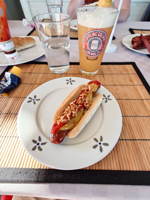 En grillkorv serverad i bröd med ketchup, senap och rostad lök på en tallrik bredvid ett glas öl och andra tillbehör på bordet.