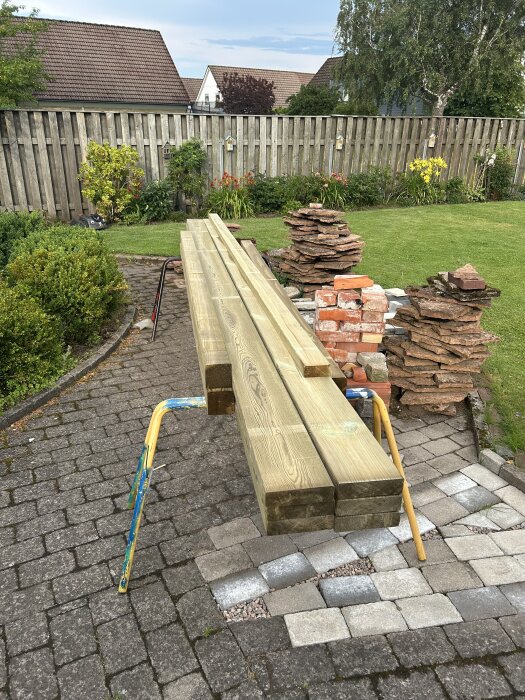 Brädor på bockar i en trädgård, omgivna av staplade tegelstenar och plattor, med en trästaket i bakgrunden.