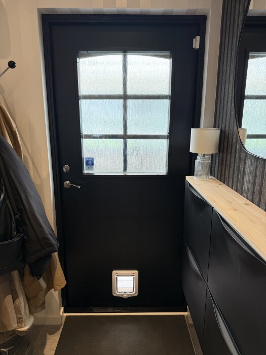 Svart ytterdörr med frostat glas i fönsterpanel och en katlucka längst ner, omgiven av förvaringsskåp och klädhängare.
