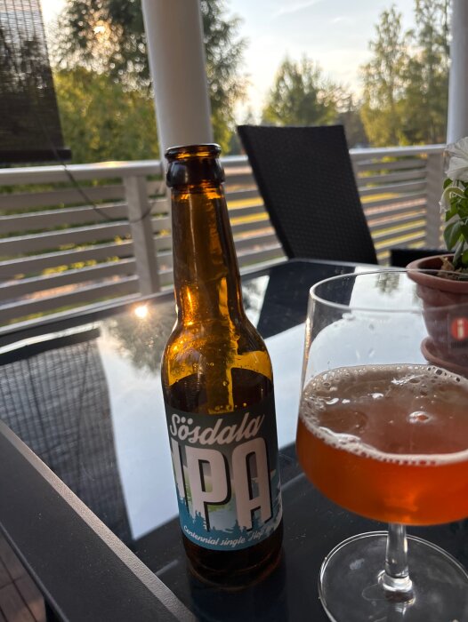 Flaska Sösdala IPA och glas med gyllene öl på glasbord utomhus, omgiven av grönska.