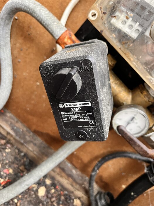 En tryckströmbrytare med inställningsknapp och en koppling till en tryckmätare syns, mot en bakgrund av flera kablar.