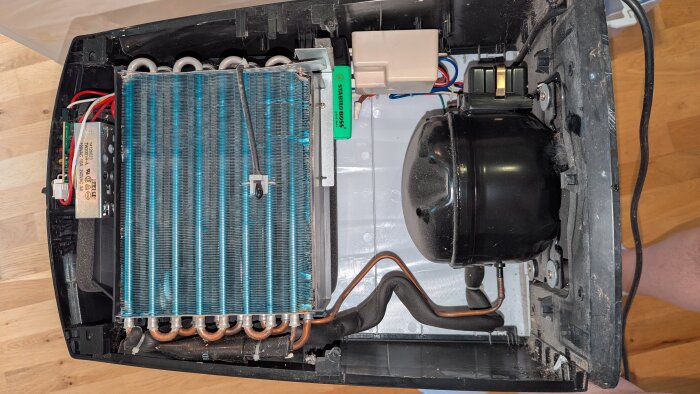 Öppet insida av en kompressor med kylslingor och kopparrör, visande en grön penna intill elektroniska komponenter och isolerade kablar.