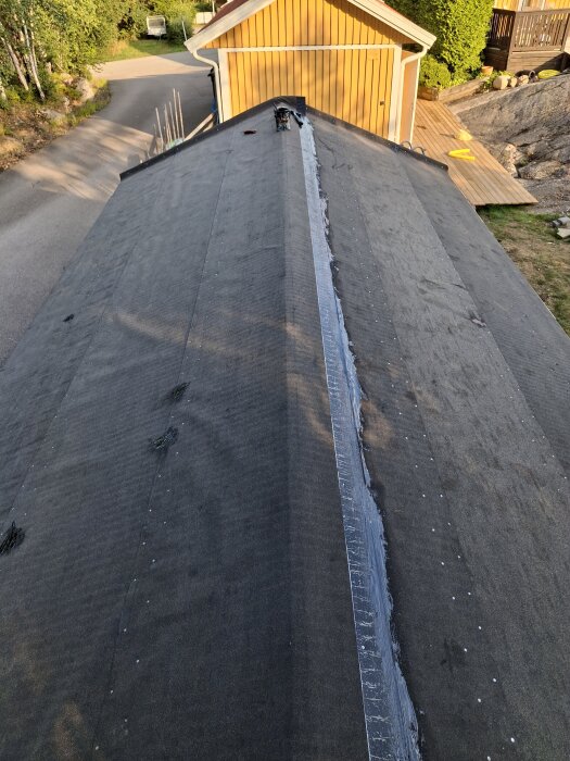 Tak med underlagspapp på friggebod, nockområde med överlapp och en trekantslist synlig längs mitten, omgiven av trädgårdslandskap och uppfart.