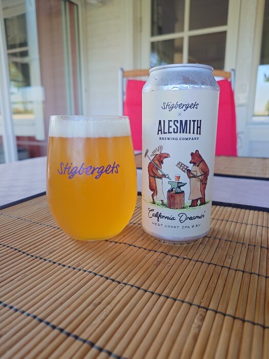 Glas med orange dryck bredvid en ölburk märkt "Stigbergets x AleSmith" på en utomhusbord med bambumatta.