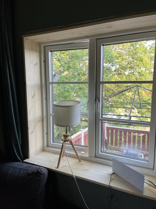 Fönster med nymonterad fönstersalning i trä, en bordslampa på fönsterbrädan, utsikt mot en trädgård med grönska och röd veranda.