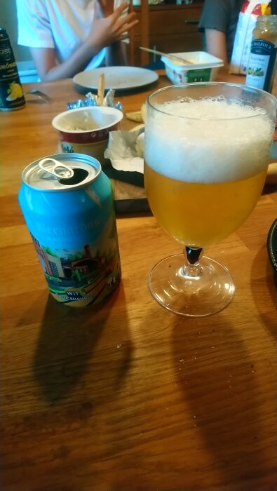 En burk St. Bernardus Wit och ett glas med belgisk wit-öl står på ett bord, omgivna av mat och dryck för en måltid.