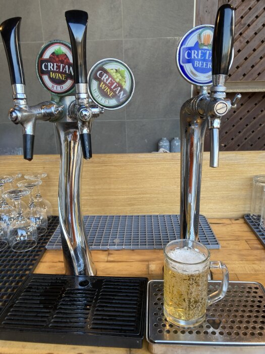 Ölpump med tre kranar märkta "Cretan Wine Red", "Cretan Wine White" och "Cretan Beer". Ett ölglas fyllt med ljus öl står på bardisken.
