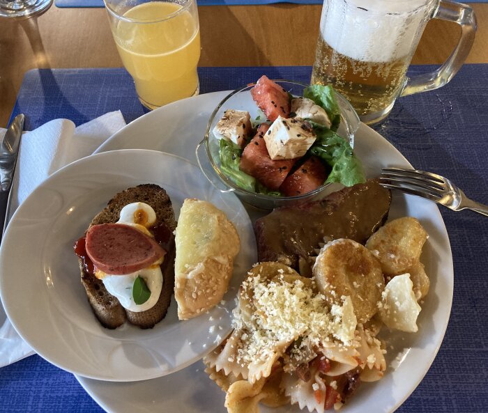 Tallrik med diverse mat: bröd med korv och ägg, pasta, potatisrätter, sallad med vattenmelon och ost. Glas med öl och juice i bakgrunden.