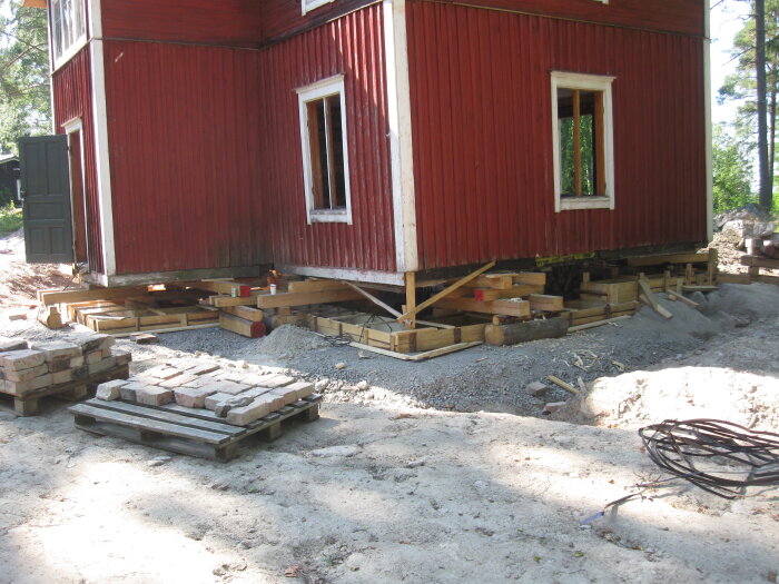 Rött hus med trästomme ovanpå betongformar och grusbädd, med lastpallar och tegelstenar i förgrunden på byggarbetsplats.