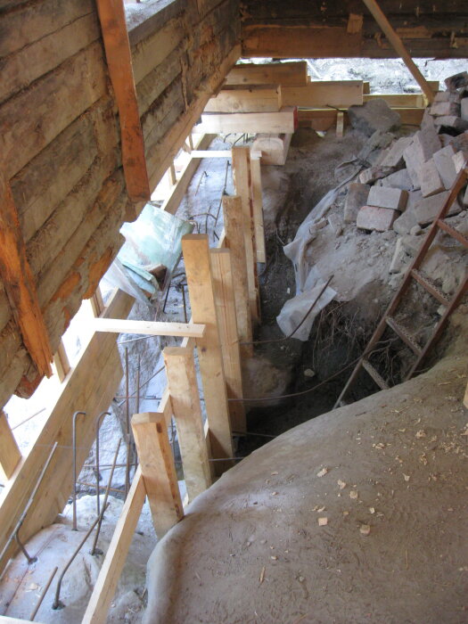 Träformar för betonggjutning, placerade på en ojämn yta nära en vägg. Sten och murbruk syns bredvid, vilket tydliggör en renovering.