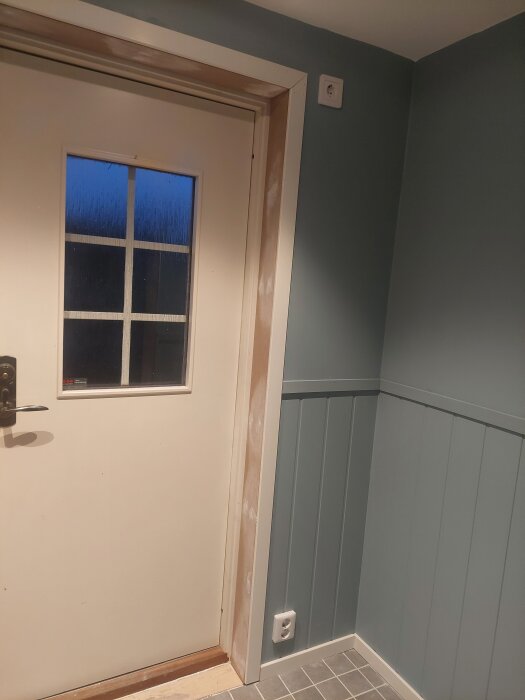 Ingång med delvis målat dörrfoder, ljus dörr med glasruta, grå väggpanel och golvsockel i rum under renovering.