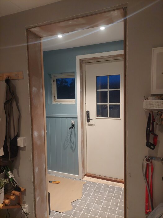 Entré med blåa väggar, vit dörr och fönster. Grovspacklade fönstersmygar, golvsocklar, och dörrfoder. Belyst med infällda spotlights. Verktyg på golvet.