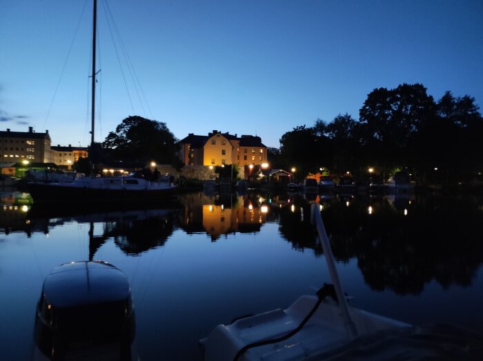 Kvällsutsikt över Bockholmssundet med båtar, upplysta byggnader och spegelblankt vatten i skymningen.