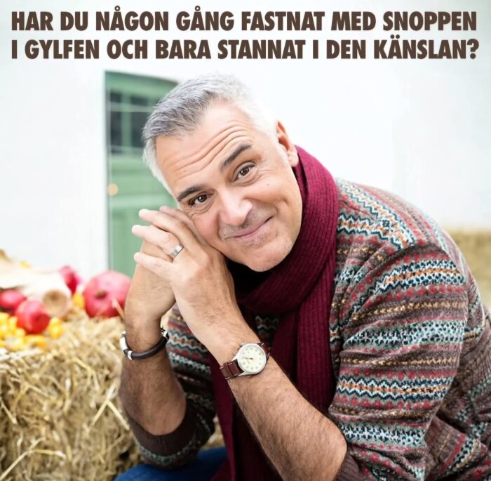 En man i mönstrad tröja och röd halsduk sitter lutad mot höloft, omgiven av frukt.