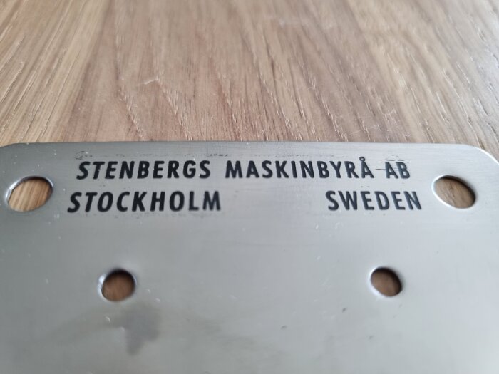 Metallskylt med texten "STENBERGS MASKINBYRÅ AB, STOCKHOLM, SWEDEN" och fyra hål, liggande på en träyta.