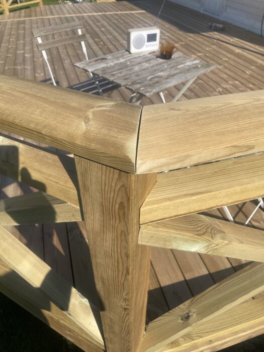 Skarv mellan två träöverliggare på altanräcke, med bord och stolar i bakgrunden på en träaltan i solljus.