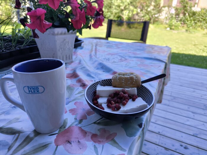 Frukostbord utomhus med kaffe, vaniljglass, smultron och en småkaka på en blommig duk i skuggan.