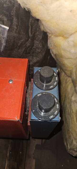 Två svarta vred med temperaturmarkeringar monterade på en äldre elpatron eller varmvattenberedare omgiven av isolering.