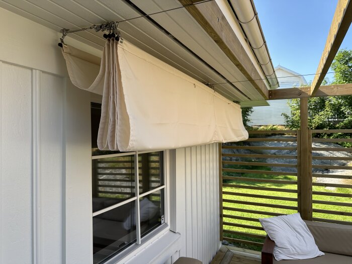 Solsegel hänger ner från taket på en pergola framför ett fönster, med trädgård i bakgrunden.