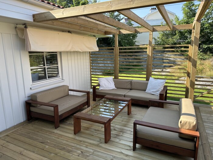 Pergola med solsegel och utemöbler mot ett hus, solsegel hänger och täcker delvis ett fönster.