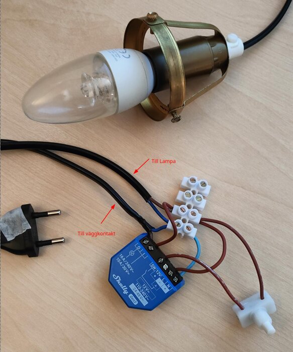 Shelly 1 gen 3 kopplad till en 5 watt LED-lampa och väggkontakt på ett skrivbord med text som markerar anslutningar.