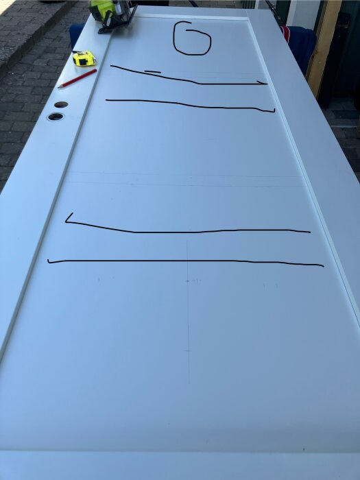 Dörr liggande på ett bord med ritade linjer och verktyg som en såg och måttband, förberett för montering av Lundbergs Orust-fönster.