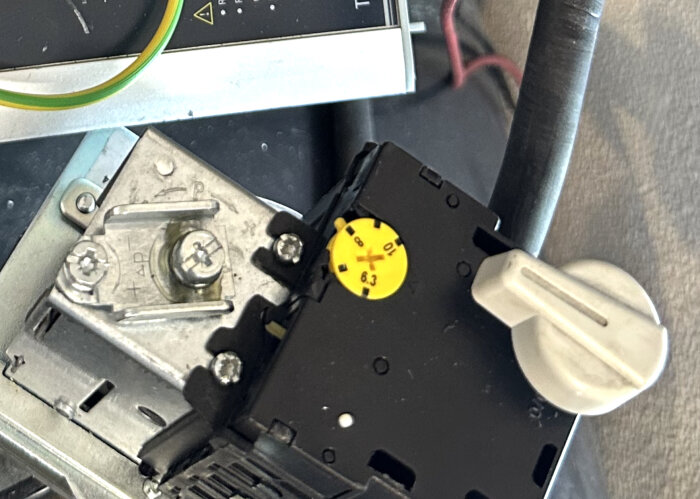 Gul ratt på svart relä med mätinställningar, bredvid vit på-av knapp och metallkomponent.