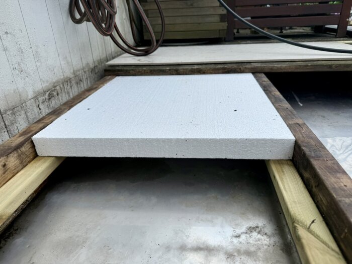 Vit cellplastskiva ligger på träbjälkar med en trätäckt yta i bakgrunden, omgiven av träramen vid byggprojekt.