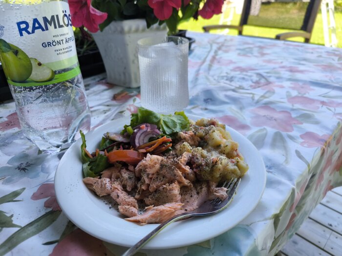 Tallrik med rökt fjällröding, potatissallad, grönsallad och Ramlösa på blommigt bord utomhus.