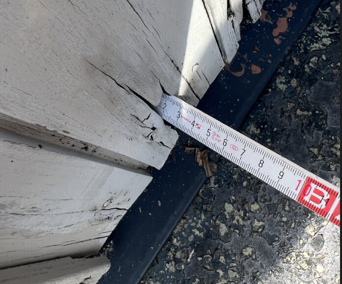 Sprucken träpanel i garagenederkant med måttstock som visar skadan, behov av renovering.