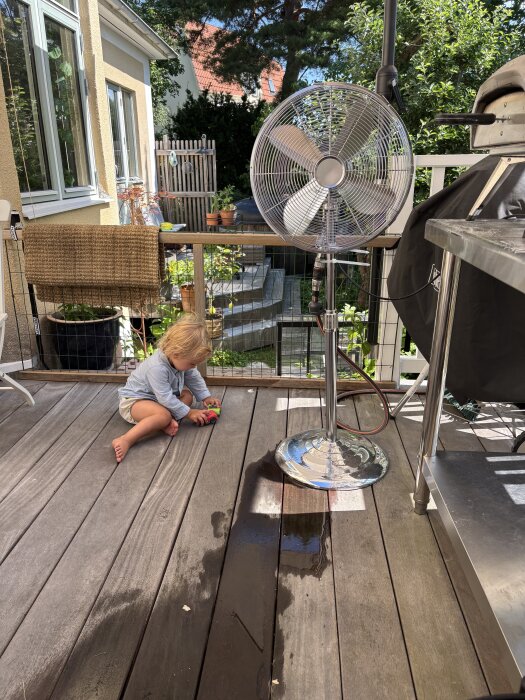 Barn leker vid foten av en stor fläkt på altanen, med vattenspridare för kylning på årets varmaste dag.