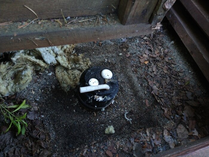 Djupborrad brunn med svart lock i marken omgiven av löv och grenar, samt något isoleringsmaterial bredvid.