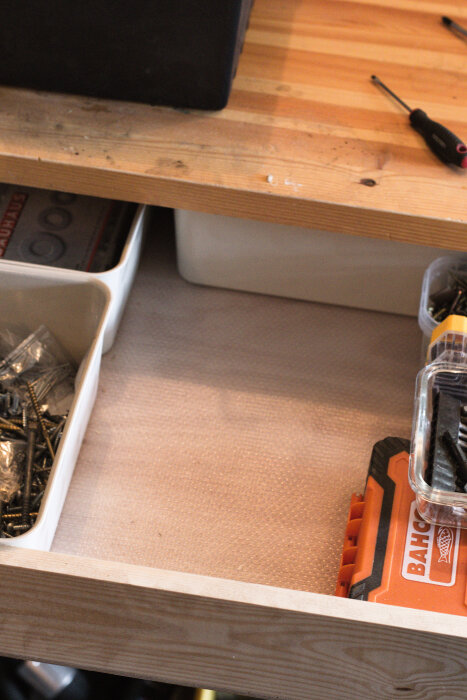 En låda med organiserade borr, sorterade i olika fack. Borr och små skruvar synliga i plast- och trälådor. Verktyg på träbänk ovanför.