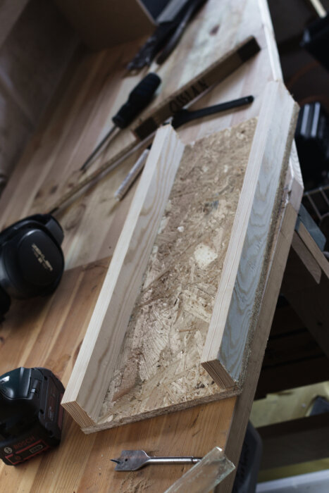 En träverktygslåda gjord av OSB-skiva och planhyvlat virke, placerad på ett arbetsbord med hörselkåpor och diverse verktyg runtomkring.