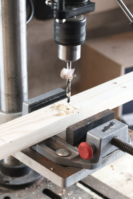 Pelarborrmaskin borrar hål i en träbit som hålls fast med ett skruvstycke, sågspån samlas runt borrhålet.