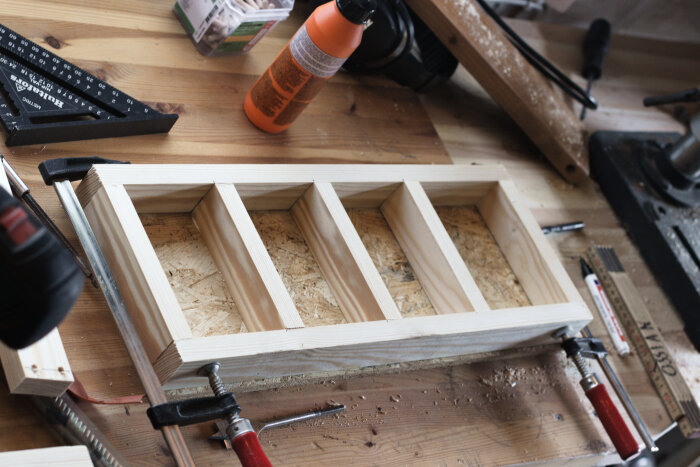 Trälåda med fyra fack, av OSB och hyvlat virke, på arbetsbänk. Verktyg och limflaska syns i bakgrunden. Lådan används för borrförvaring.