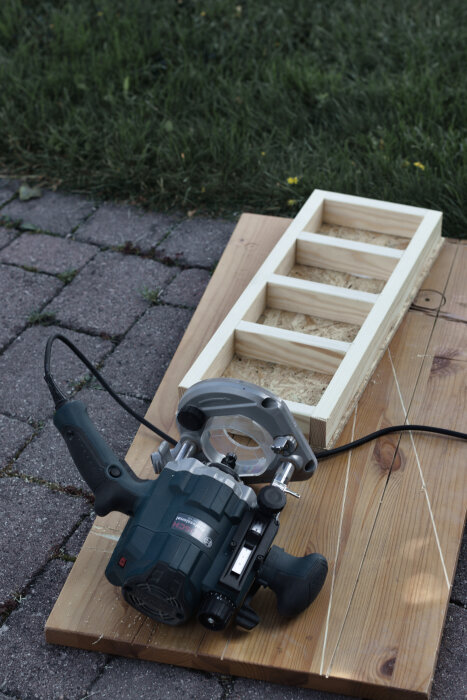 Träkonstruktion för borrförvaring på en träplatta med en fräs placerad bredvid, liggande på stenbelagd yta.