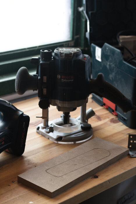 En Bosch-handöverfräs på ett arbetsbord med en MDF-bit med konturer ritade, för förberedelse av hylla till en minidammsugare.