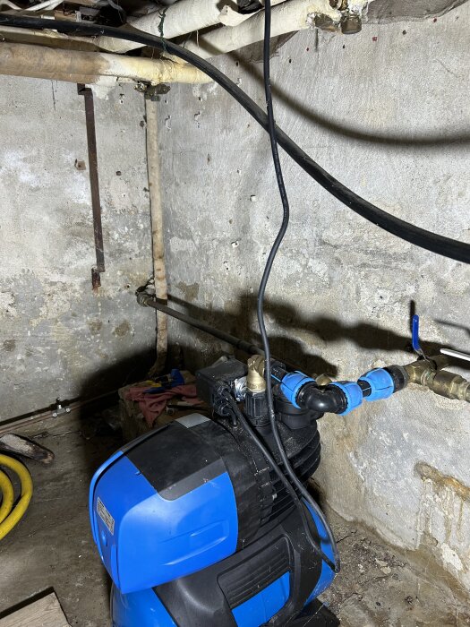 Blå dränkbar länspump med röranslutning i hörnet av källarvägg, omgiven av kablar och andra ledningar.
