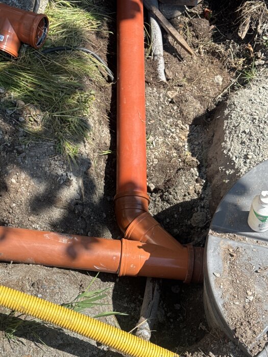 Orange avloppsrör med ett Y-böj i marken istället för ett T-böj, omgiven av gräs och jord.