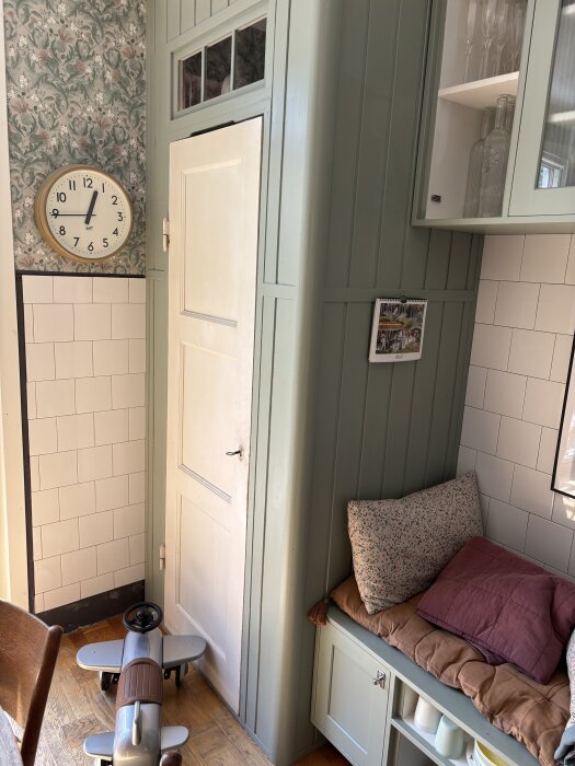 Grönmålad vägg med inbyggd djup skåpdörr, väggklocka ovanför, leksaksflygplan på golvet, bänk med kuddar bredvid.