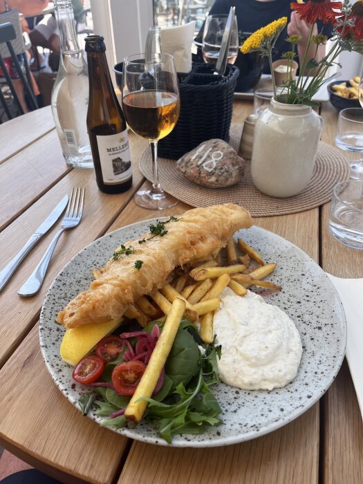 Tallrik med fish and chips, grönsaker, citron och sås på ett träbord med dryck i glas.
