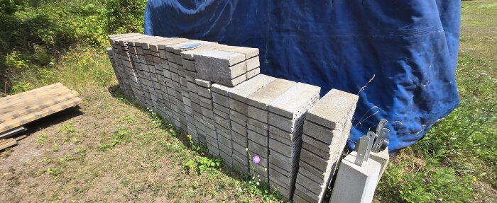 Grå betongblock staplade på varandra utomhus, delvis täckta av en blå presenning, omgivna av gräs och några blommor.